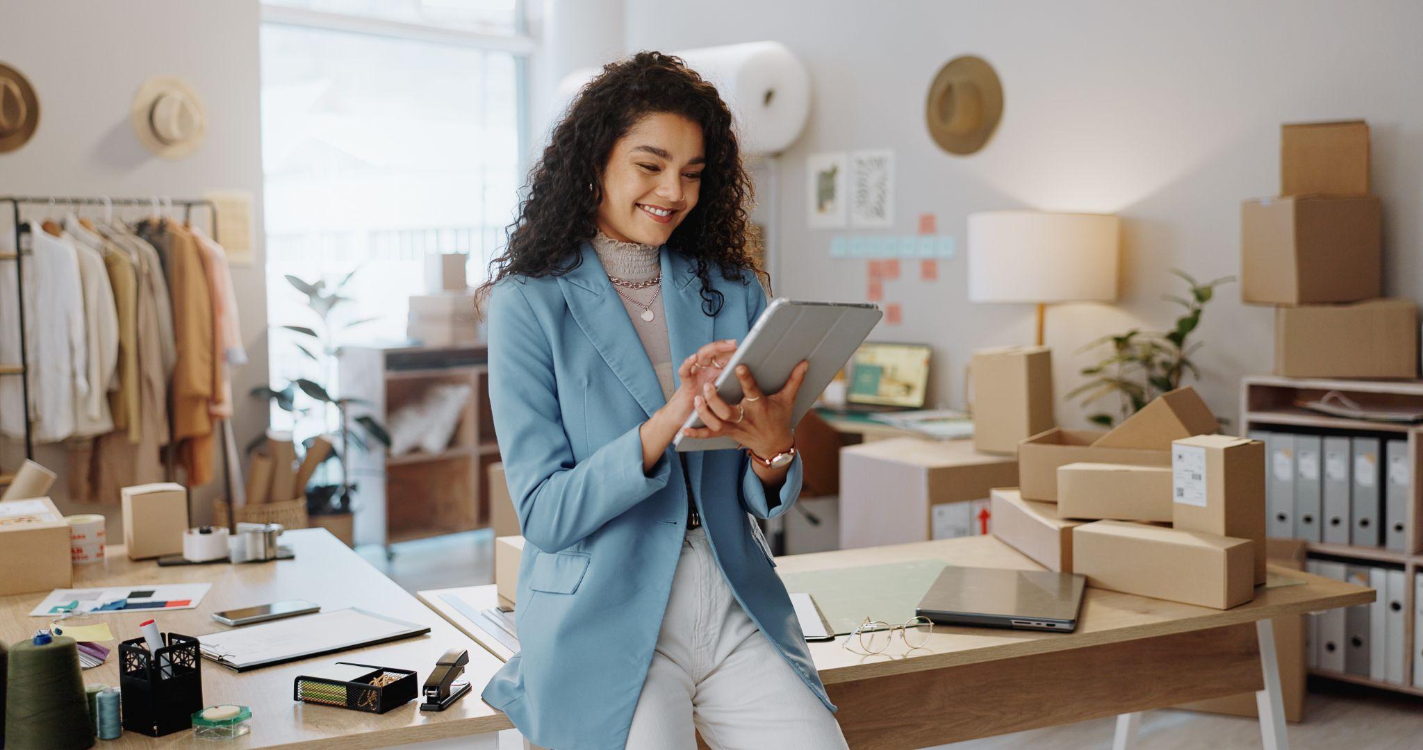 Commerçante dans son atelier, tenant une tablette, cartons et matériel d’expédition derrière — préparation des commandes pour la boutique.
