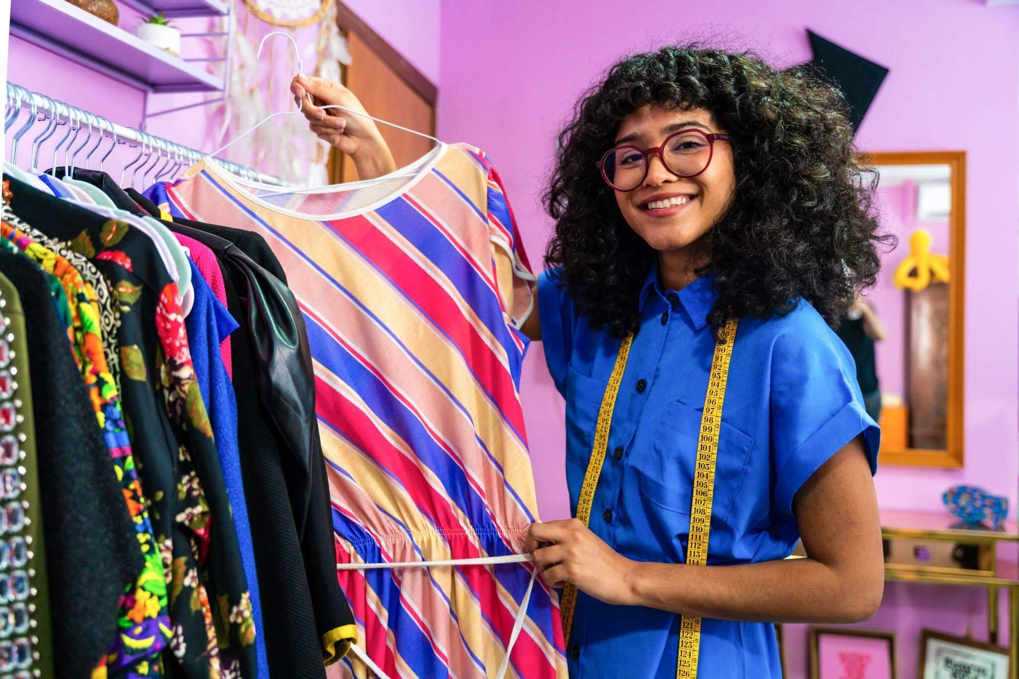 Costurera sonriente con una cinta métrica alrededor del cuello presentando una pieza de su colección.