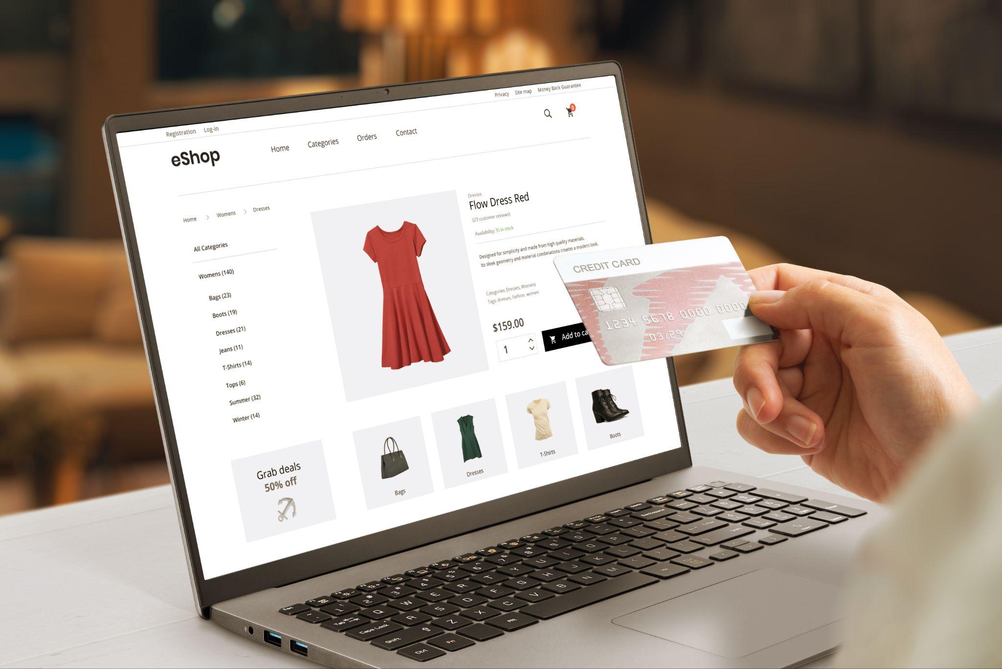 Schermo del computer che mostra una scheda prodotto per un abito rosso, mano che tiene una carta bancaria - acquisto da una boutique di moda online.
