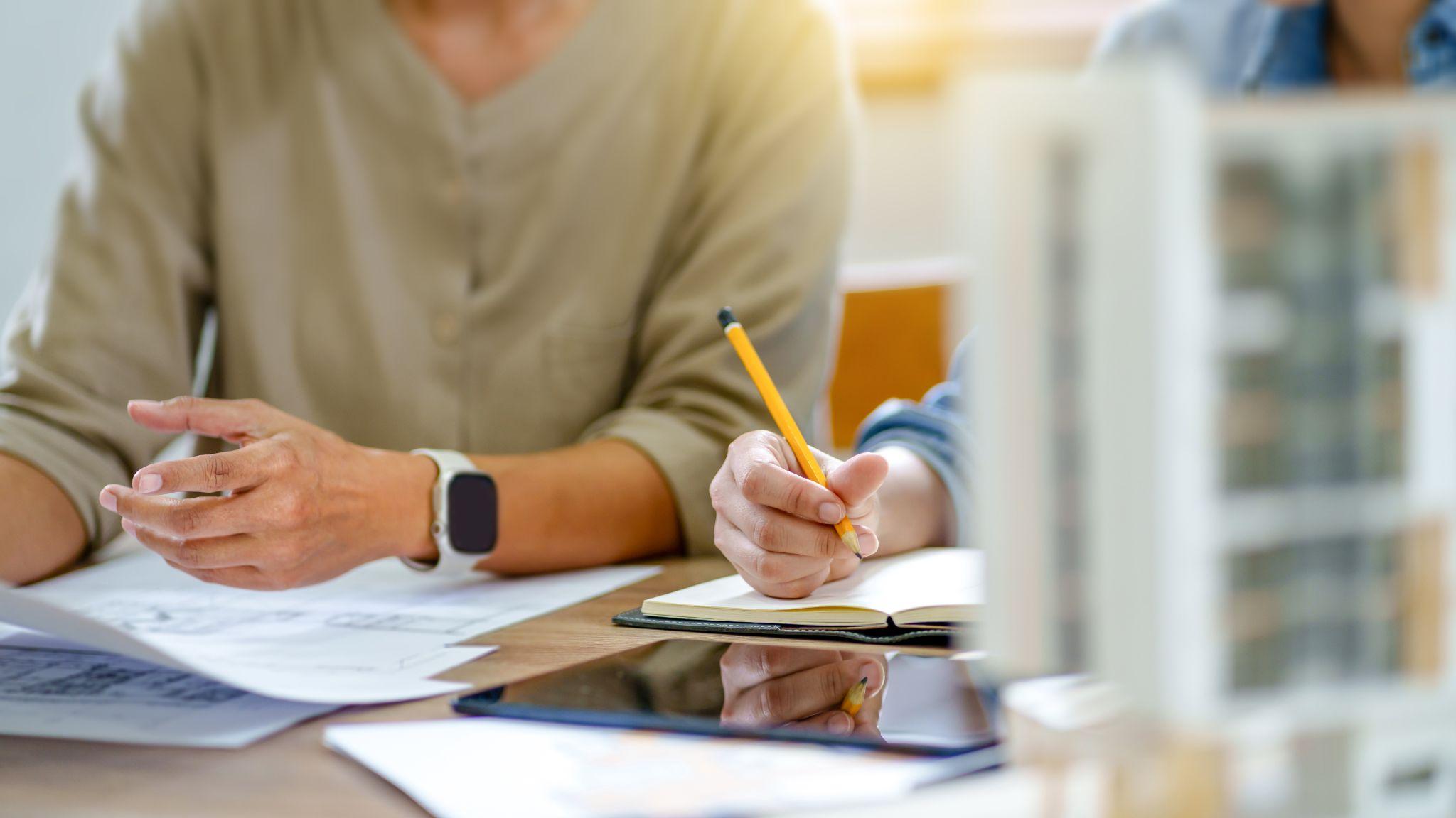 Due persone in riunione sono sedute a un tavolo. Una ha in mano un foglio di carta, l'altra sta scrivendo su un quaderno.