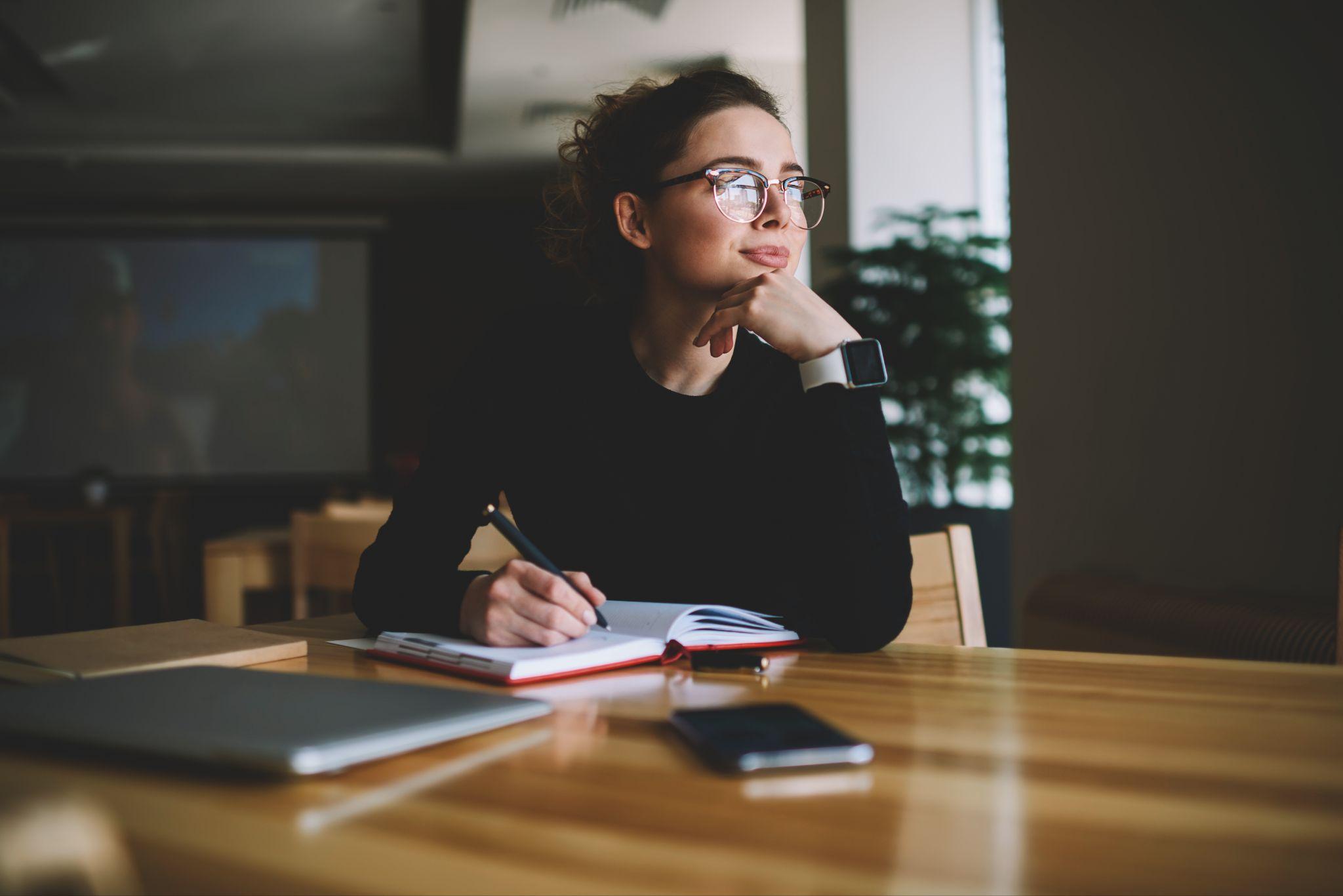 Femme entrepreneuse réfléchissant et écrivant dans un carnet, ordinateur portable sur la table d’un café — préparation du sourcing grossiste mode.