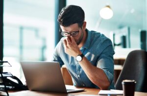 Homme entrepreneur penché sur son bureau, main au front, carnet et ordinateur portable allumé — réflexion avant l’ouverture d’une boutique de mode / prêt-à-porter (sourcing)