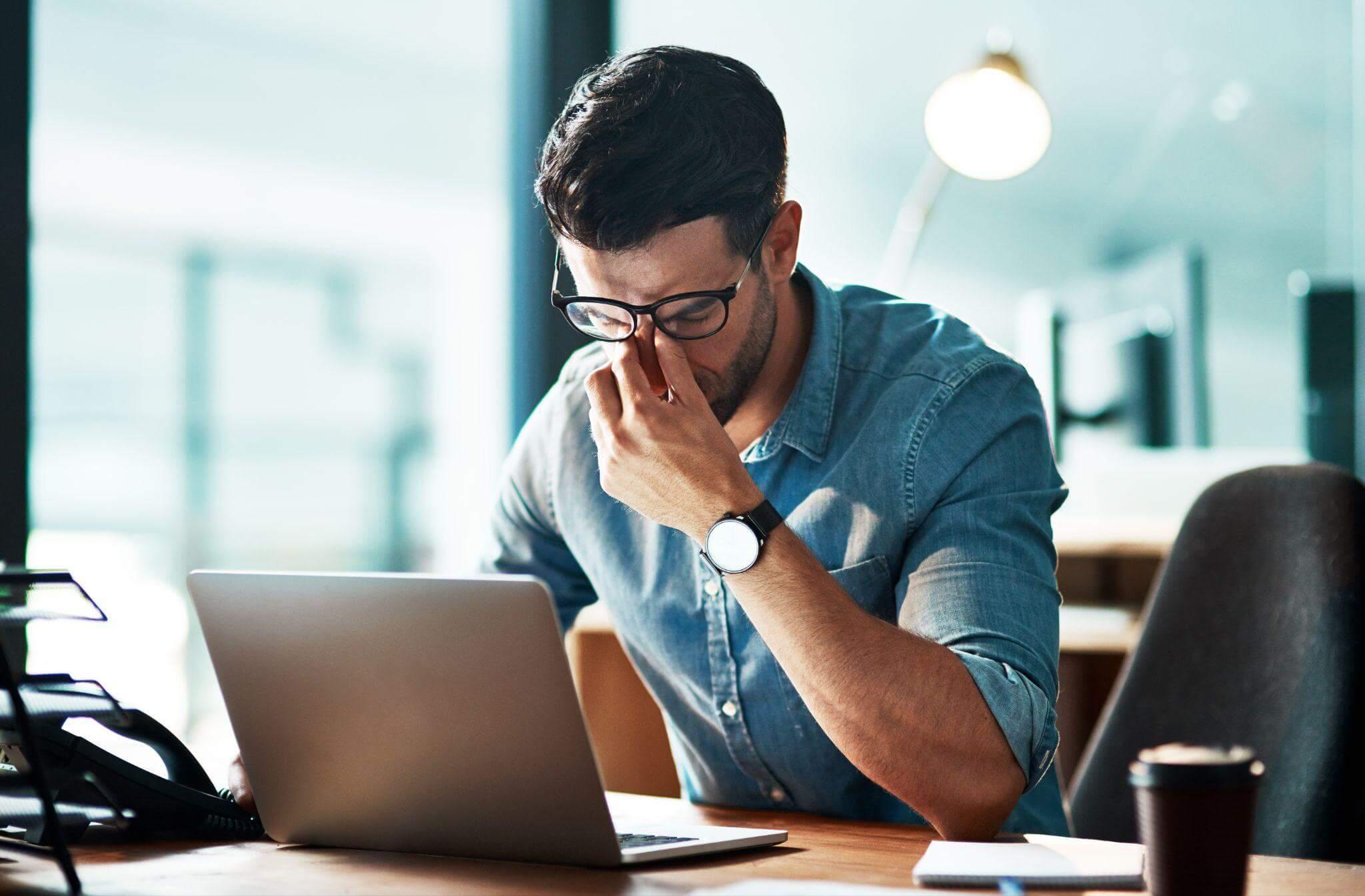 Homme entrepreneur penché sur son bureau, main au front, carnet et ordinateur portable allumé — réflexion avant l’ouverture d’une boutique de mode / prêt-à-porter (sourcing)