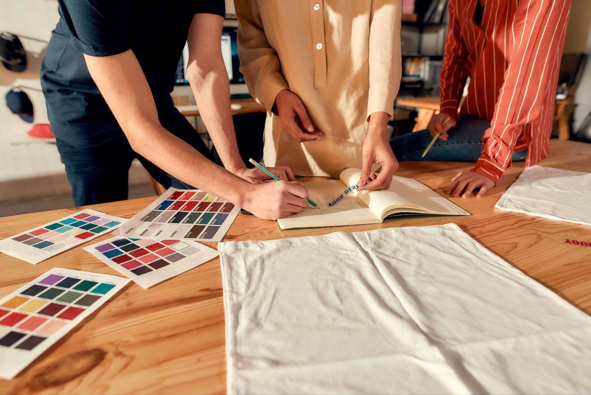 Três pessoas trabalham com materiais, têxteis e cores à volta de uma mesa.