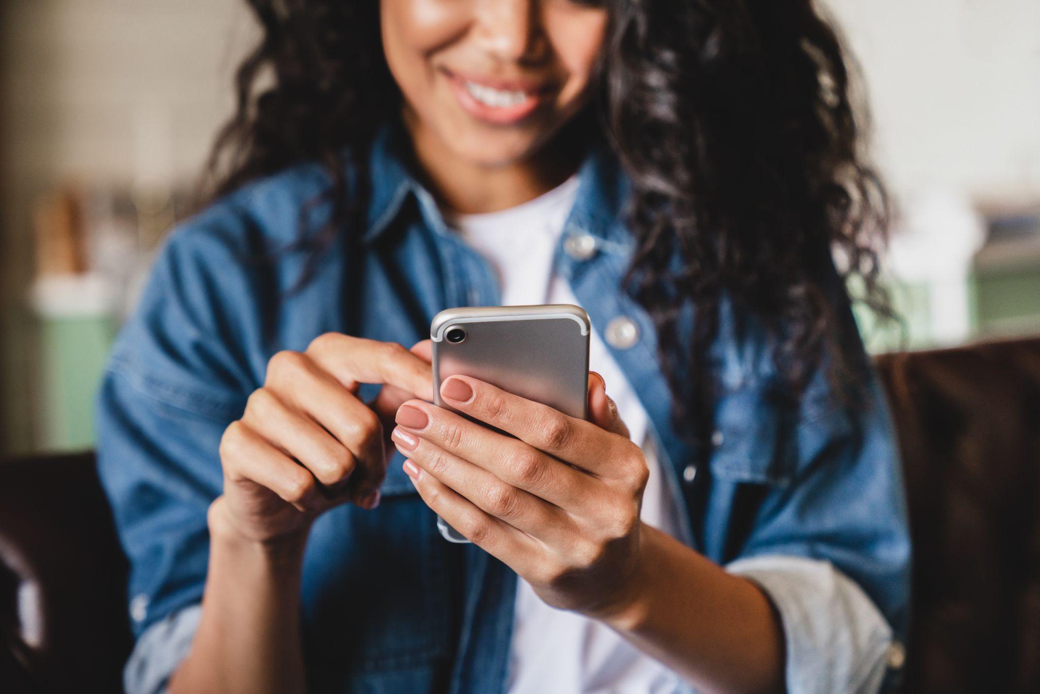 Une femme assise et souriante scroll sur son smartphone avec sa main droite. Elle utilise whatsapp business pour vendre plus simplement.