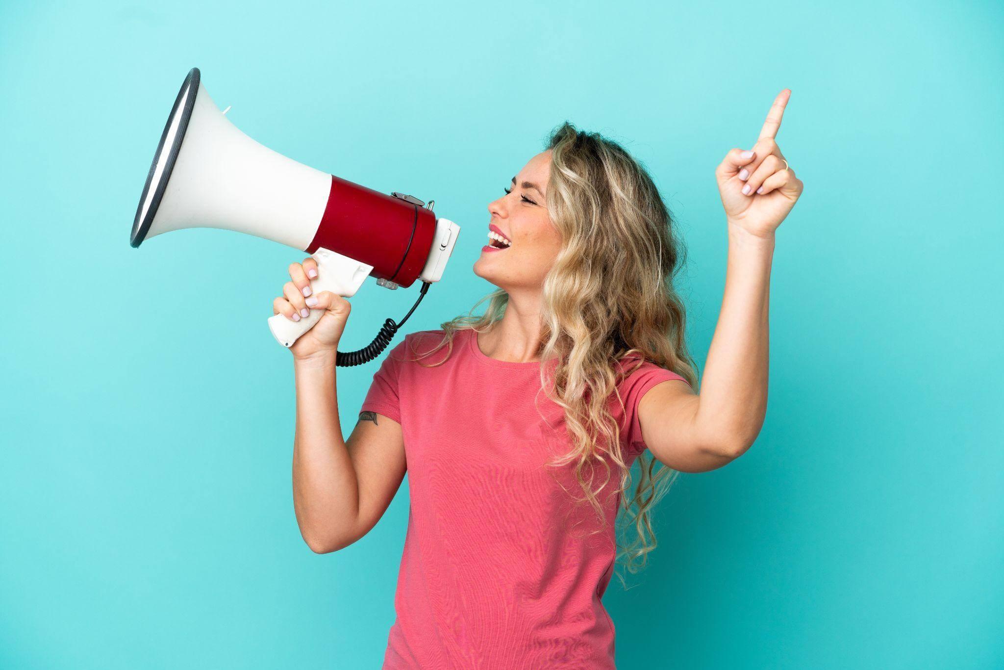 Une femme avec un t shirt rose devant un fond bleu tient un mégaphone pour tester son concept auprès de prospects.