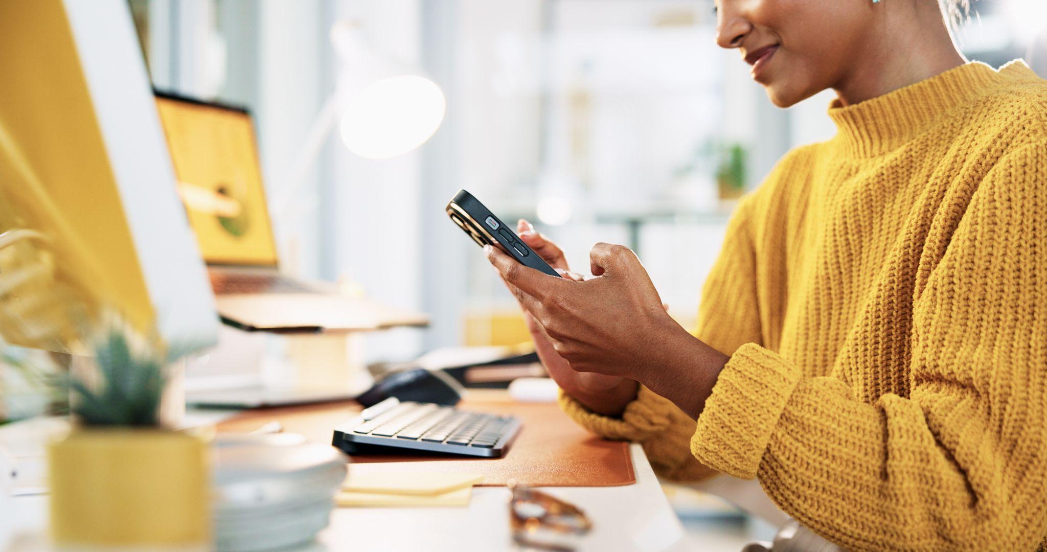 Une femme est assise à son bureau devant ses écrans et son ordinateur. Elle tient et travaille ses newsletters avec son smartphone.