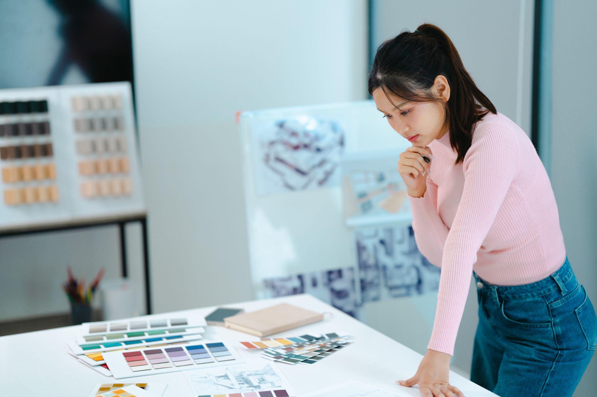 Une femme est debout devant une table et réfléchit à ses créations en fonction de sa cible. Elle regarde des palettes de couleurs et différents textiles.