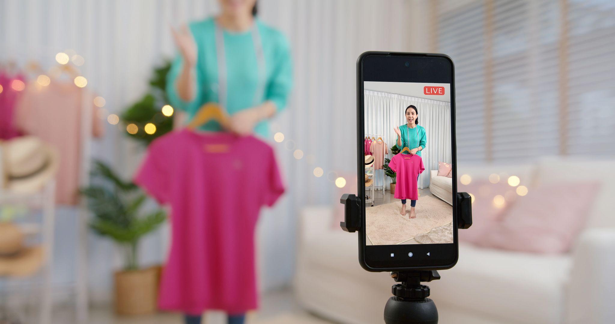 Une femme présente un haut rose en live avec son smartphone posé sur un trépied.