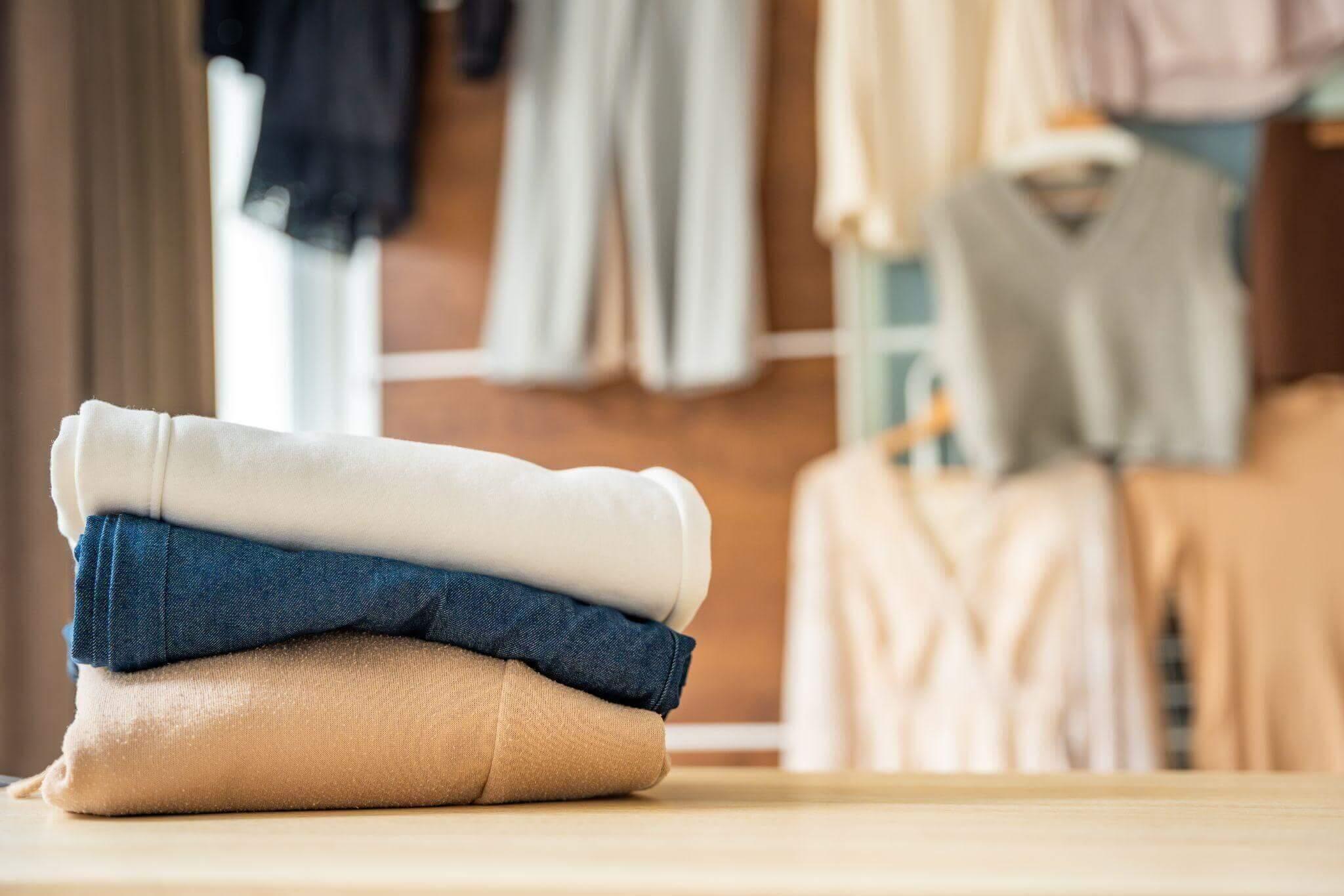 Uma pilha de três peças de vestuário está em cima de uma mesa. No fundo desfocado, um quarto de vestir com roupas igualmente brilhantes e feitas de matérias-primas de qualidade. 